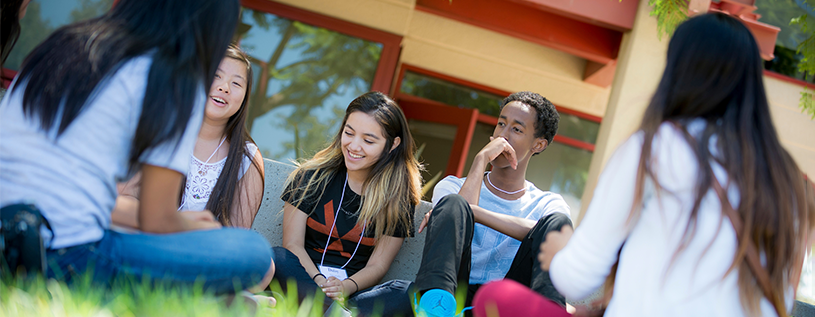 Five students sitting in green grass in a circle smiling and talking